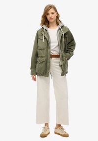 Olivgrön arbetsjacka med huva, dragsnören och framfickor, kombinerad med vita vida byxor och ljusa sneakers.