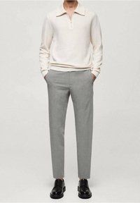 Homme portant un pull col zip à manches longues beige clair, un pantalon gris slim et des chaussures habillées en cuir noir, les mains dans les poches.