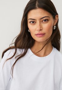 Camiseta blanca de algodón con escote redondo, corte relajado y mangas cortas; modelo que lleva pendientes de aro grandes de plata.