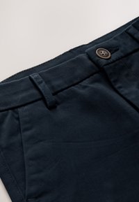 Gros plan d'un pantalon bleu marine foncé montrant une taille boutonnée, une boucle de ceinture et une poche avant sur fond clair.
