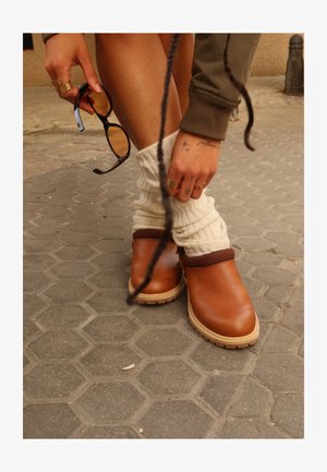 Persoană purtând pantofi maro slip-on și încălzitoare albe pentru picioare, ținând ochelari de soare negri, cu tatuaje vizibile pe mâini și inele, pe o pavăză de piatră.