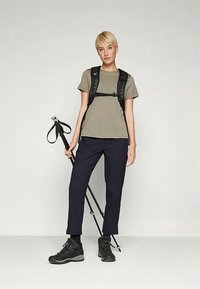 Žygio apranga, sudaryta iš alyvuogių žalios spalvos marškinėlių, juodo taktinio kuprio, tamsiai mėlyno kelnių ir juodų žygių batų, laikant trekingo lazdą.