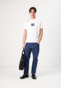 White cotton t-shirt featuring a blue CK logo, paired with blue denim jeans. Model holds a black leather tote bag. Black shoes complete the outfit.