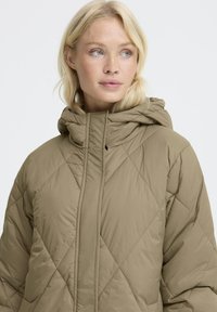 Blonde Frau, die eine gesteppte, beige Daunenjacke mit hohem Kragen und Kapuze trägt und leicht zur Seite blickt, vor einem schlichten Hintergrund.