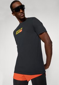 Black short-sleeve t-shirt with "BERLIN RUN" in yellow and a red box. Worn with orange shorts layered over black tights. Black sunglasses.