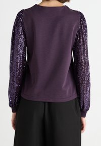 Sudadera púrpura con mangas de lentejuelas texturizadas, escote redondo y corte relajado, combinada con pantalones negros.