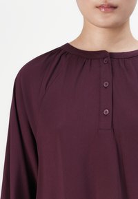 Person wearing a dark purple blouse with a round neckline and three buttons on the front placket, shown from neck to shoulder.