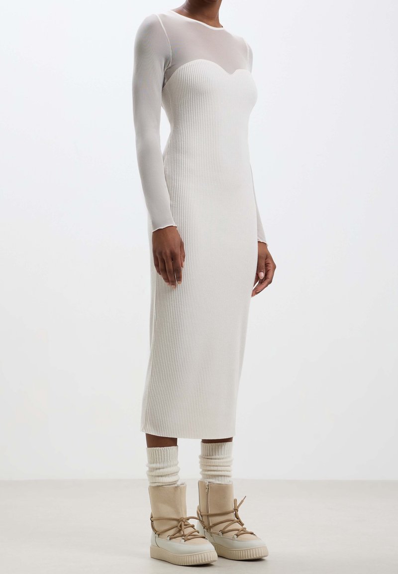 Robe blanche à côtes avec des manches longues et un corps transparent. Associée à des bottes crème jusqu'au genou et des chaussettes en tricot texturé. Design simple et ajusté.
