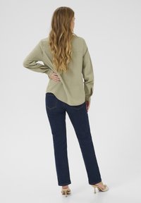 Licht olijfgroene blouse met lange mouwen en een gebogen zoom, gecombineerd met donkerblauwe skinny jeans en beige hakken, bekeken vanuit de achterkant.