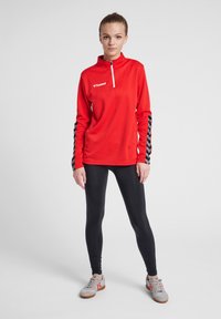 Maglia atletica rossa a maniche lunghe con colletto con zip, tessuto testurizzato e strisce nere ai lati. Abbinata a leggings neri e scarpe da ginnastica grigio chiaro.