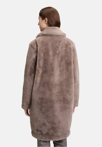 Kvinde iført en lang, blød gråbrun faux fur frakke set fra ryggen, kombineret med ternede bukser mod en ensfarvet hvid baggrund.
