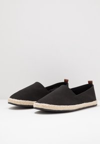 Black canvas slip-on shoes with a rounded toe, jute rope sole, and small brown leather accents on the heel. Lightweight and flat design.