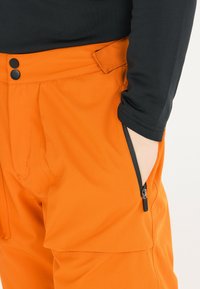 Orangefarbene Hose aus leichtem Stoff, mit einer seitlichen Reißverschlusstasche, zwei schwarzen Knöpfen am Bund und einer glatten Textur.