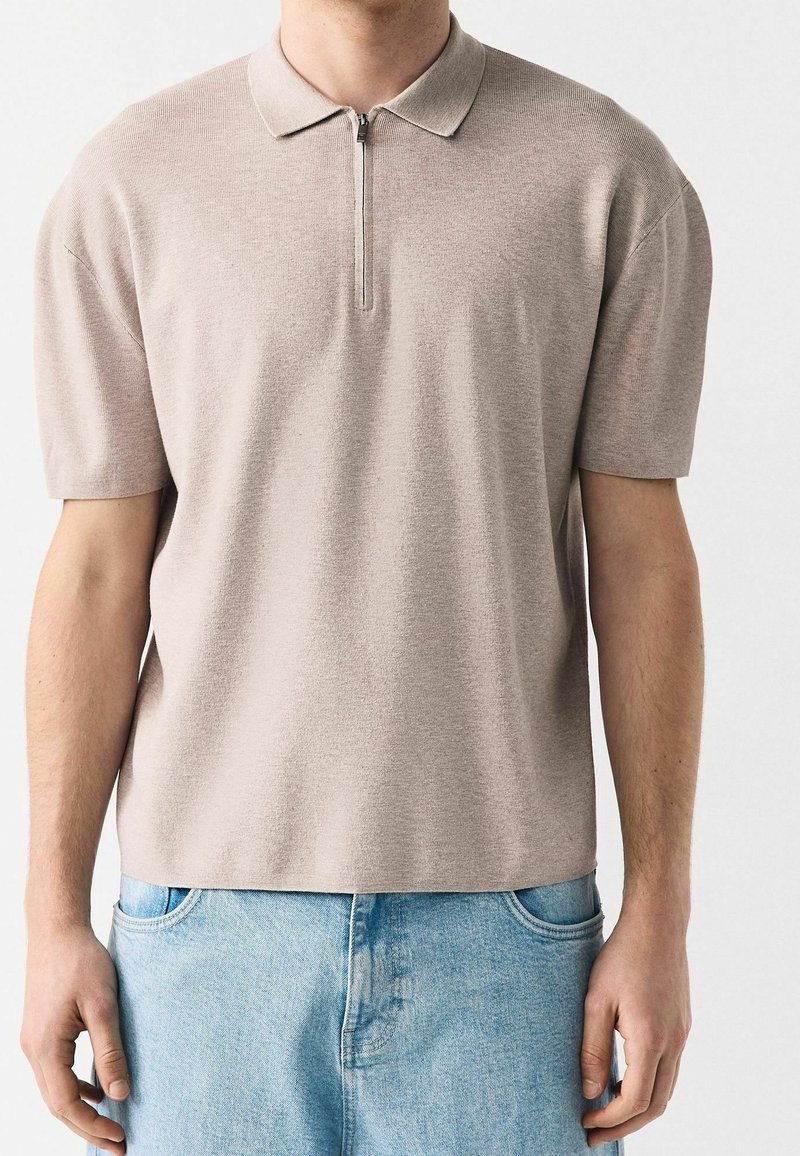 Homme portant un polo à manches courtes beige clair avec une fermeture éclair à l'avant et un jean bleu clair, debout contre un fond uni.
