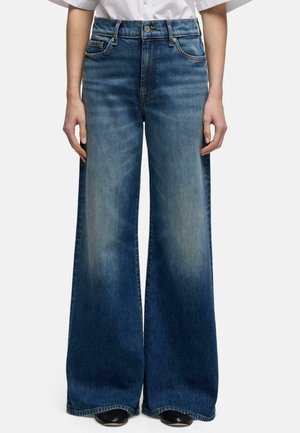 Personne portant un jean bleu taille haute à jambes larges avec des panneaux avant délavés, associé à des chaussures noires et une chemise blanche.