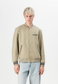 Beige wool-blend varsity jacket with button closure, side pockets, and black accent stripes on cuffs. Features embroidered logo on chest.