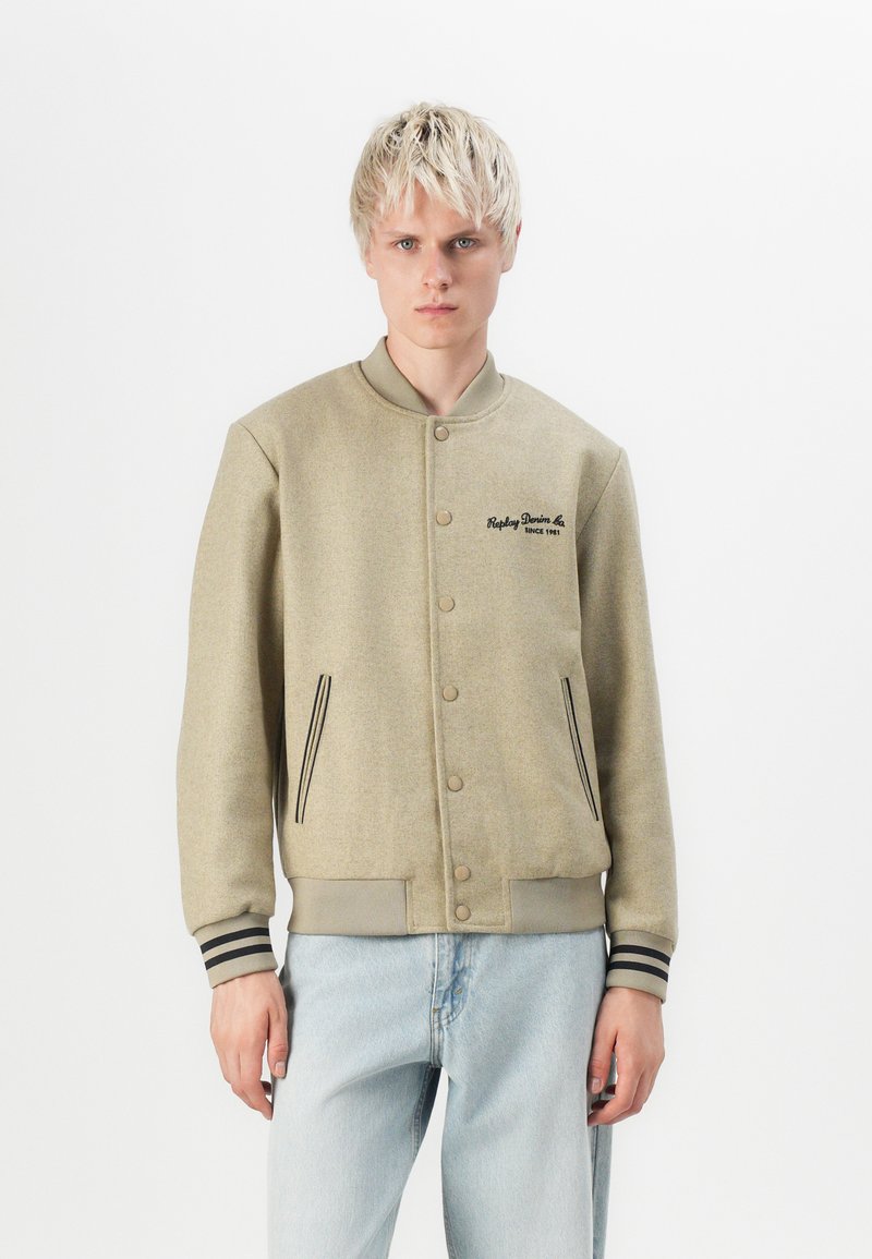 Beige villasegu segu jacket, millel on nööbid ja küljetaskud, ning mustad aktsenttriibud varrukatel. Rinnal on brodüüritud logo.