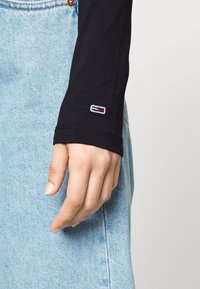 Svart långärmad topp med en tajt passform, med en liten broderad logotyp i blått, rött och vitt nära ärmslutet, tillsammans med ljusblå jeans.