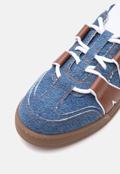 Κοντινή λήψη ενός sneaker από τζιν με λευκά κορδόνια, καφέ δερμάτινες λεπτομέρειες και καφέ λαστιχένια σόλα σε λευκό φόντο.