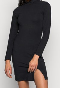 Czarna sukienka z długim rękawem, wysokim dekoltem, dopasowanym krojem i rozcięciem do uda. Wykonana z gładkiego, elastycznego materiału.