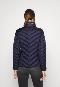 Kvinna med brunt hår i hästsvans som bär en figursydd marinblå vadderad jacka och mörkgrå jeans, stående med ryggen mot kameran.