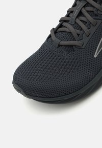 Chaussure de sport noire avec une tige en mesh respirant, surface texturée et embout renforcé. Comprend des accents gris et des lacets plats.