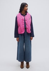 Ροζ γιλέκο από φλις με στρογγυλή λαιμόκοψη, κουμπιά μπροστά και τσέπες, φορεμένο πάνω από μπλε τζιν μπλούζα και φαρδιά τζιν παντελόνια. Καφέ μποτάκια μέχρι τον αστράγαλο.