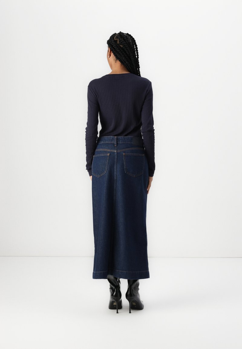 Marineblaues geripptes Langarmoberteil, kombiniert mit einem dunkelblauen Jeansmaxirock. Der Rock hat eine hohe Taille, zwei Gesäßtaschen und einen geraden Schnitt. Schwarze Stiefel.