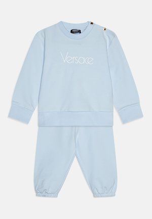 Tuta in cotone azzurro chiaro composta da una felpa con colletto a girocollo, logata "Versace" in ricamo e pantaloni con polsini elasticizzati. Accenti in ottone dorato.