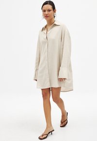 Abito camicia in lino beige con colletto, chiusura a bottoni e maniche oversized. Indossato con sandali con tacco marroni.