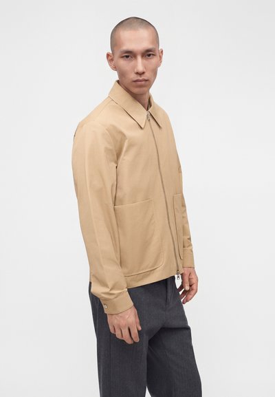 Homme aux cheveux courts portant une veste beige zippée avec de grandes poches avant et un pantalon gris foncé, debout sur un fond blanc uni.