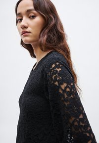 Μαύρο τοπ με δαντέλα και μακριά, διαφανή μανίκια που διαθέτουν πολύπλοκα λουλουδάτα σχέδια· στρογγυλεμένος λαιμός και υφή υφάσματος που αναδεικνύουν το σχέδιο.