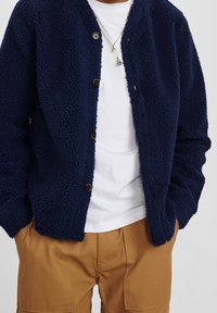 Marinblå fleecekofta med texturerat, fluffigt material. Har en rund halsringning, framknappar och en avslappnad passform. Bärs över en vit t-shirt.