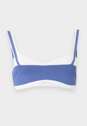 Hvit bikinioverdel med bred horisontal blå stripe over fronten og blå stropper på ensfarget lyst bakgrunn.