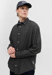 Camisa de cuadros en blanco y negro, de botones y de manga larga, con un corte entallado y detalles en el dobladillo lateral, acompañada de una gorra oscura.