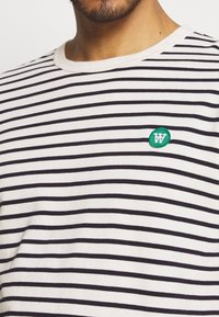 Randig t-shirt i gräddvit och marinblå, med rund halsringning och en grön broderad logotyp. Tyget verkar mjukt och något strukturerat.