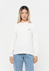 Hvid langærmet bomulds T-shirt med rund hals og lille Carhartt-logo i orange på venstre bryst, sammen med lys blå stribede jeans.