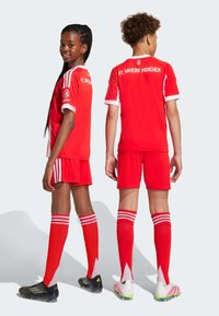 Červený futbalový dres a šortky FC Bayern Mníchov s bielymi akcentmi, spojené s červenými ponožkami s bielymi pruhmi a čiernymi a dúhovými kopačkami.