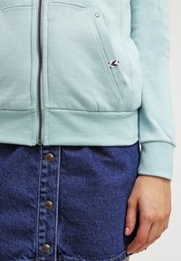 Felpa zip-up azzurro chiaro con tessuto testurizzato e tasche laterali, abbinata a una gonna in denim blu con chiusura a bottoni e una finitura liscia.