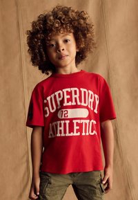 Rød bomulds-T-shirt med teksten "SUPERDRY ATHLETIC" i hvid. Korte ærmer og afslappet pasform. Sammen med olivengrønne cargo shorts.