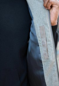 Sivi kaput s teksturiranom vanjštinom i glatkom unutrašnjošću, s mekanim detaljem i patentnim zatvaračem na lijevoj strani. Crna unutrašnjost je vidljiva.