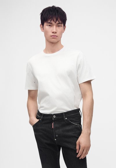 Jeune homme aux cheveux courts et noirs portant un t-shirt blanc uni et un jean noir, debout avec une main dans la poche sur fond blanc.