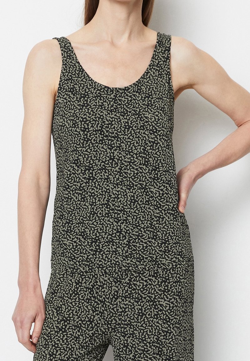 Femme portant un haut sans manches noir et gris à motifs et un pantalon assorti, main posée sur la hanche, debout devant un fond blanc.