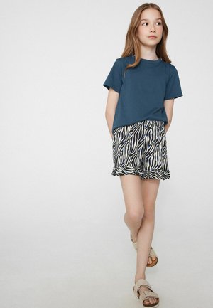 Niña con una camiseta azul de manga corta, pantalones cortos con estampado de cebra y sandalias beige, de pie con las manos detrás de la espalda sobre un fondo blanco.