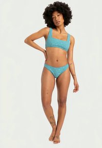 Τηλεκατευθυνόμενο μπικίνι σε τιρκουάζ χρώμα με τετράγωνη λαιμόκοψη και λεπτές τιράντες πάνω. Χαμηλοκάβαλο κάτω μέρος. Διαθέτει υφή με μοτίβο σε όλη την επιφάνεια.