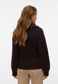 Hrubě pletený svetr tmavě hnědé barvy s vysokým límcem, žebrovanou strukturou a širokými rukávy, doplněný světle béžovými kalhotami.