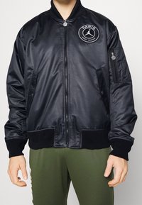 Black bomber jacket made of glossy fabric, features a round embroidered logo on the chest, zipper pockets, ribbed cuffs, and hem.