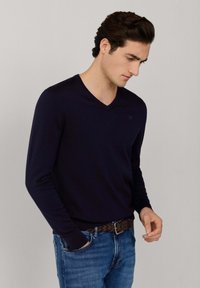 Mann trägt einen dunkelblauen V-Ausschnitt-Pullover und blaue Jeans mit geflochtenem braunem Gürtel, steht mit einer Hand in der Tasche vor einfarbigem Hintergrund.