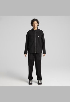 Zwarte zip-up jack met twee borstzakken en logo, gecombineerd met bijpassende zwarte broek met grote voorkruimelzakken, beide in een relaxte pasvorm.