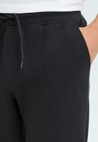 Ciemnoszare, teksturowane spodnie dresowe z elastycznym pasem i sznurkiem. Posiadają boczne kieszenie oraz małą czarną naszywkę z logo na udzie.